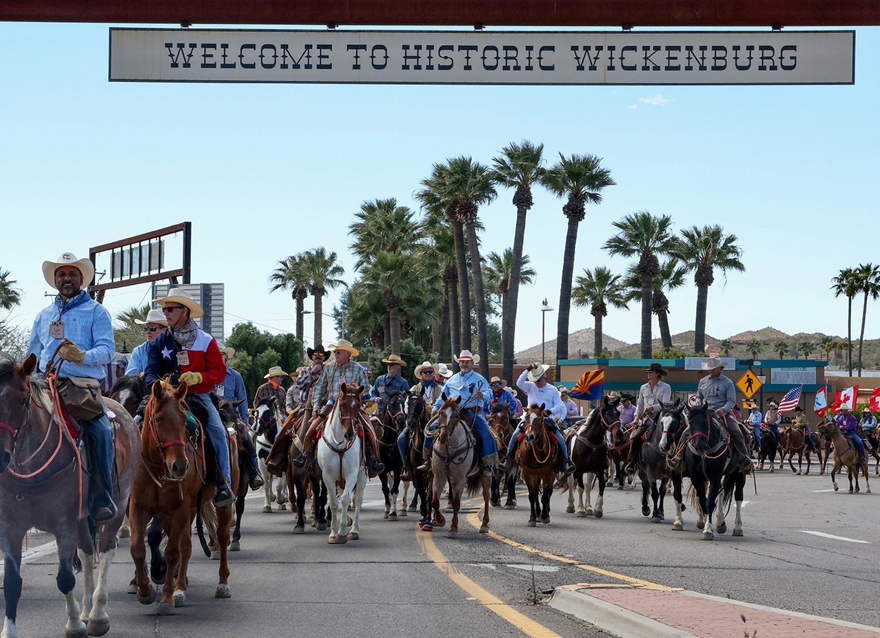 Riders on horseback parade under the Welcome to Historic Wickenburg sign.